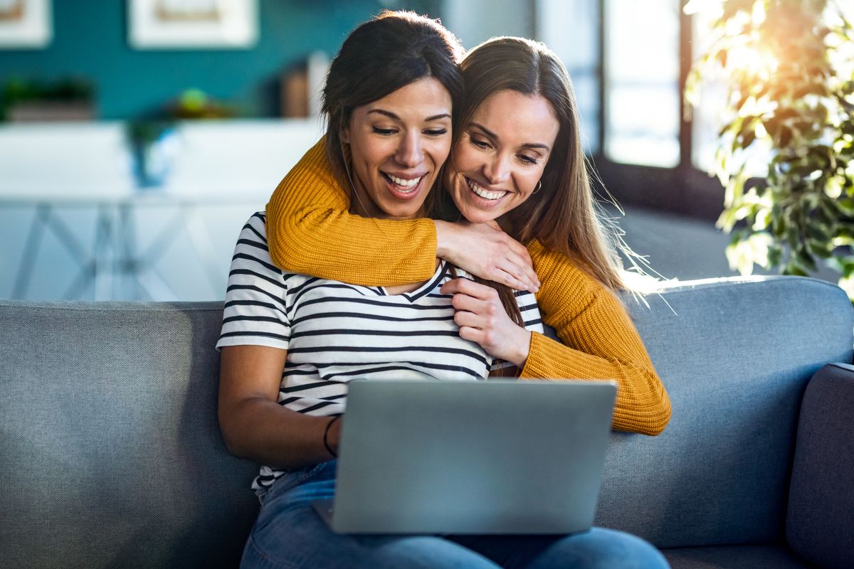 Two women smiling together at a laptop on a sofa
