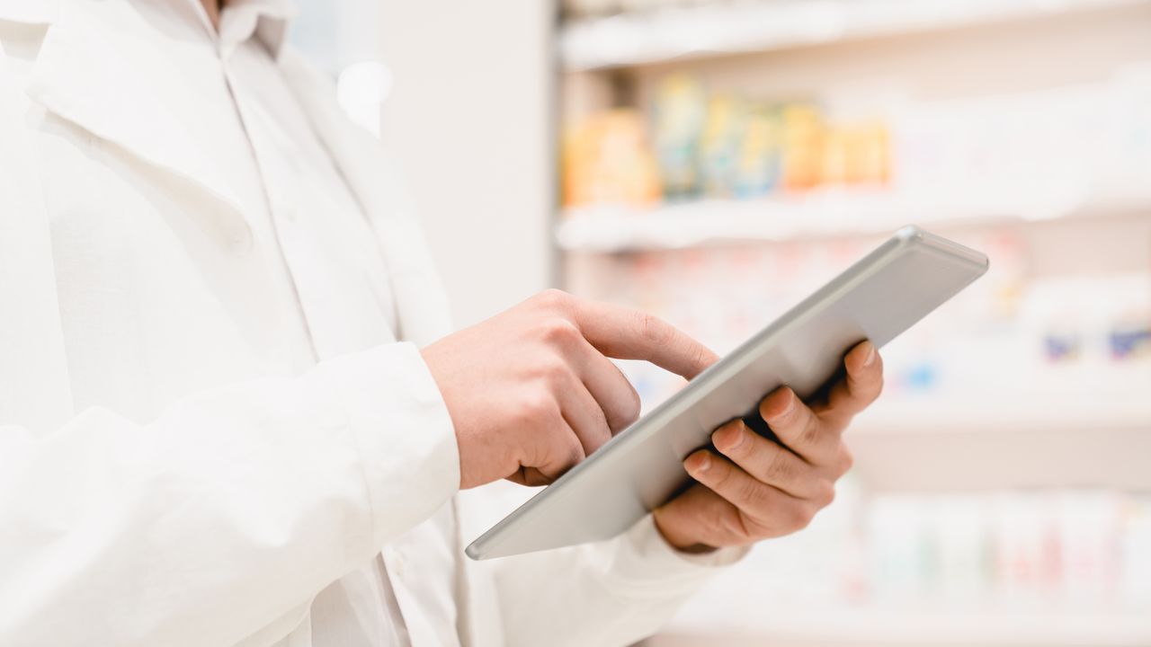 A pharmacist using a tablet in front of stocked pharmacy shelves
