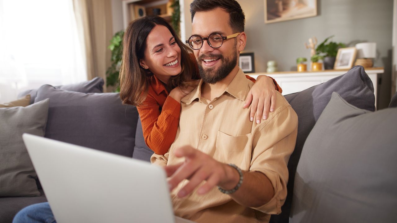 A couple smiling together at a laptop on their sofa